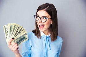 Cheerful businesswoman holding bills of dollar over gray background. Wearing in blue shirt and glasses.
