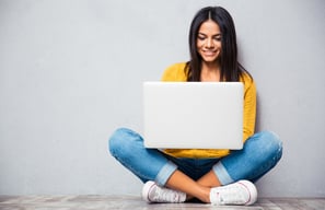Happy young woman sitting on the floor with crossed legs and using laptop on gray background.jpeg